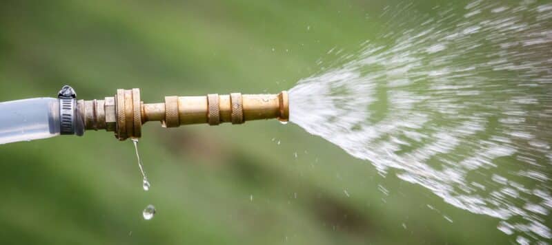 A brass water hose nozzle sprays a steady stream of water with droplets trailing behind against a blurred green background